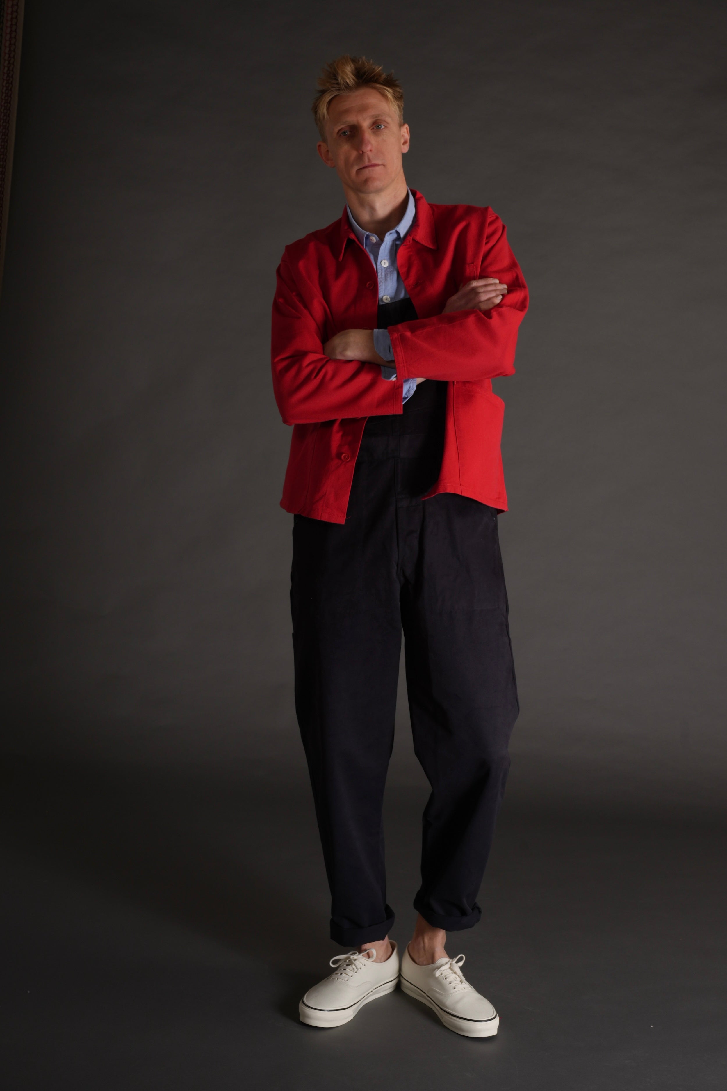 Tommy wears Carrier Company red garment washed cotton work jacket over navy blue dungarees and pale blue chambray shirt with no socks and white plimsoles