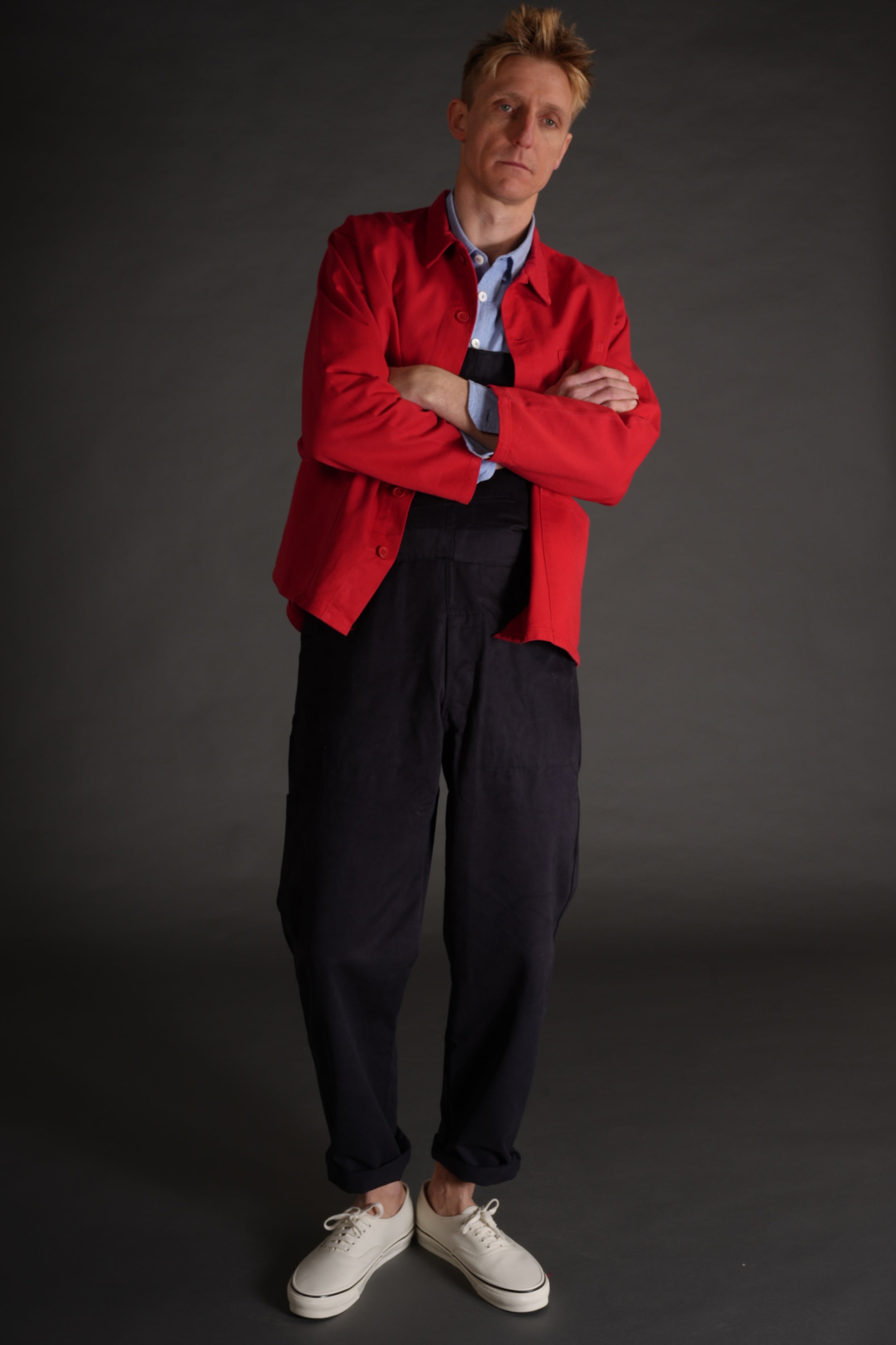 Tommy wears Carrier Company red garment washed cotton work jacket over navy blue dungarees and pale blue chambray shirt with no socks and white plimsoles