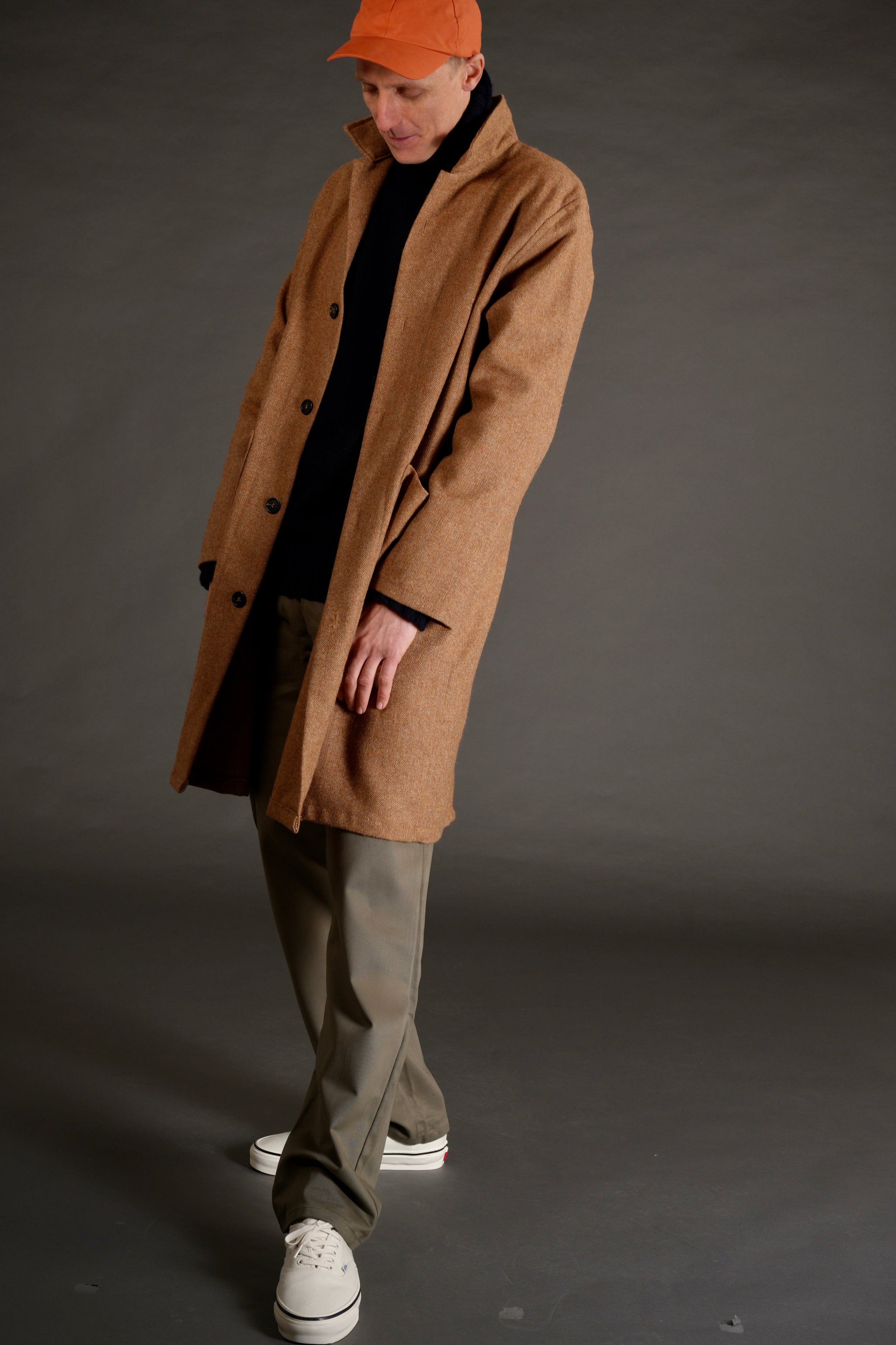 Tommy wears tan herringbone wool coat over navy roll neck with olive mens work trouser and orange waxed cap