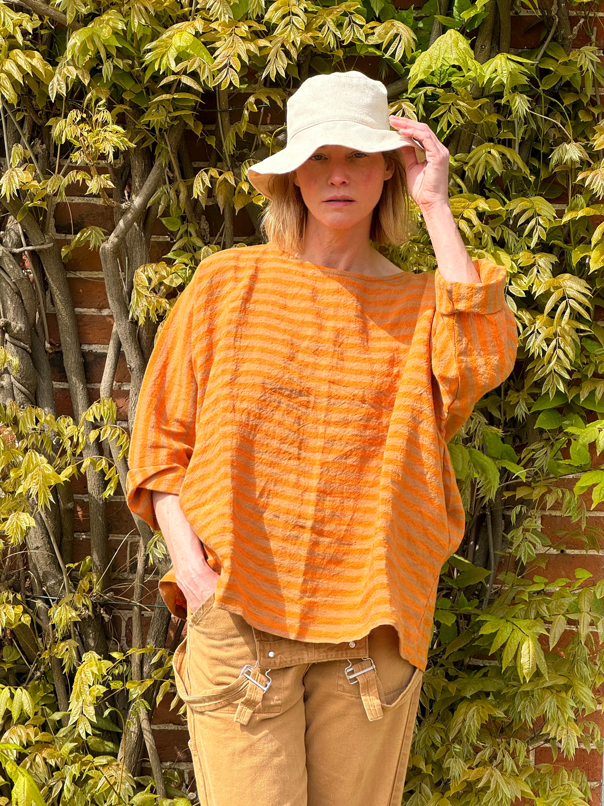 Sienna wears Orange and tan high necked boxy top with pockets and 3/4 length sleeve, with tan dungarees and sun hat