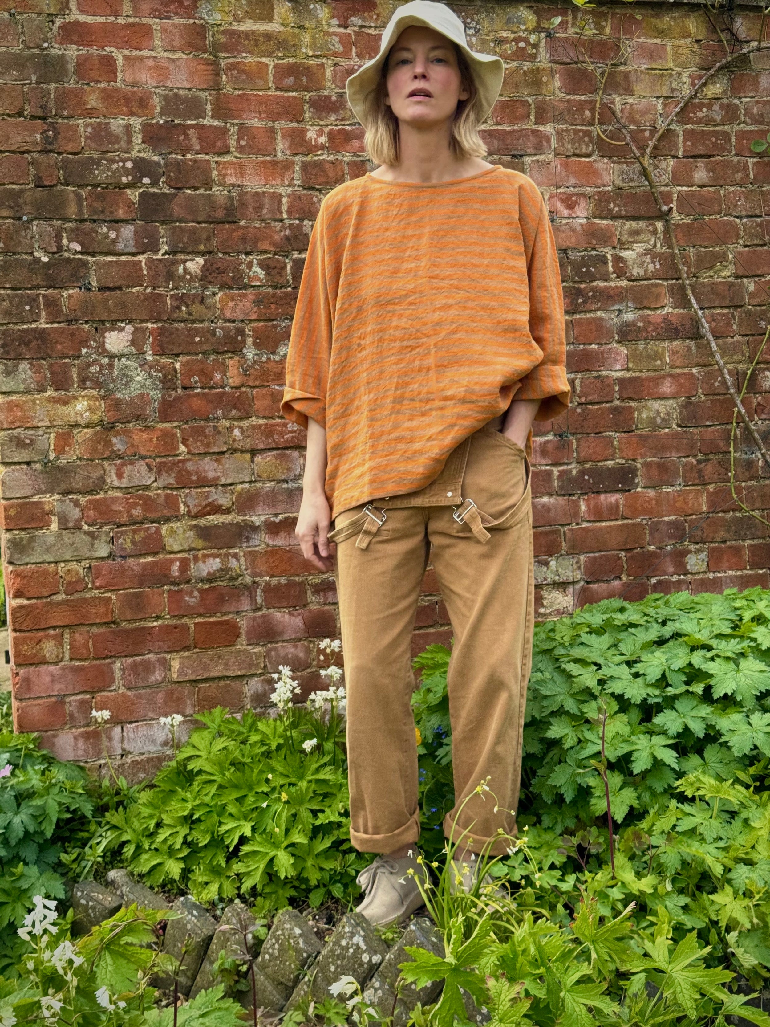Sienna wears Orange and tan high necked boxy top with pockets and 3/4 length sleeve, with tan dungarees and sun hat
