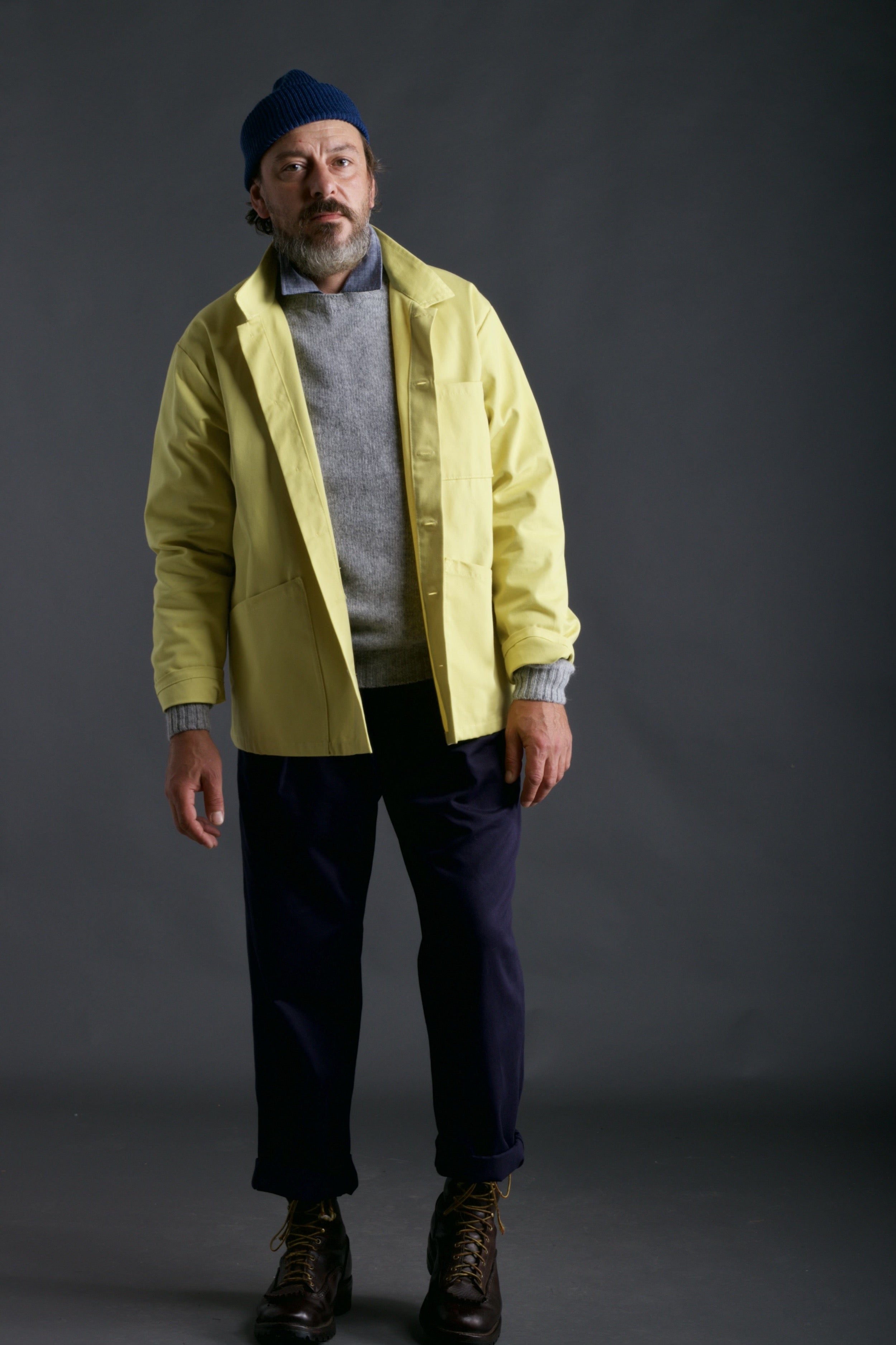 Man wears Carrier Company Classic Trouser in Navy Drill with Chambray Shirt, Shetland Lambswool Jumper in Silver, Work jacket in Citron and Wool Hat in Petrol Blue