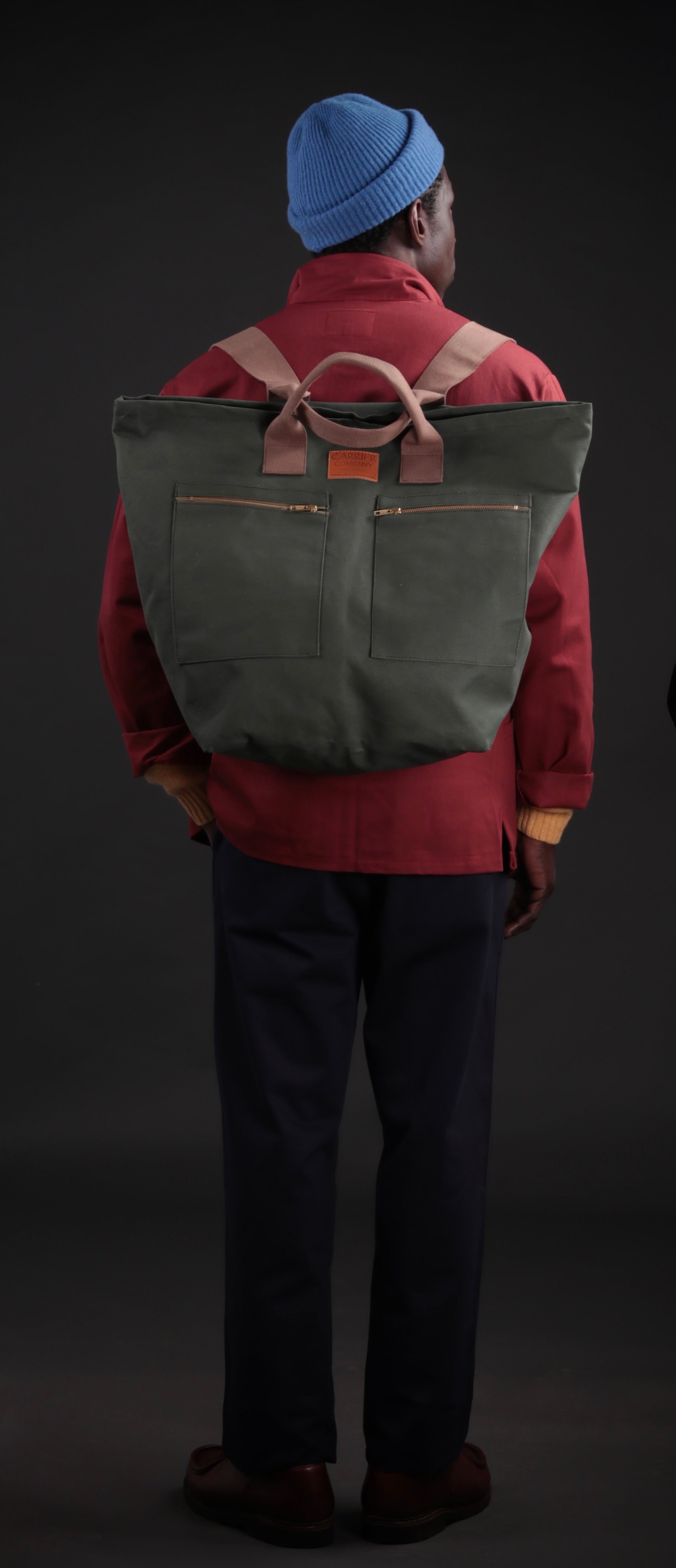 Man carries Carrier Company Backpack in Olive. Wearing Norfolk Work Jacket in Breton, Navy Classic Trousers and a Turquoise Donegal Hat