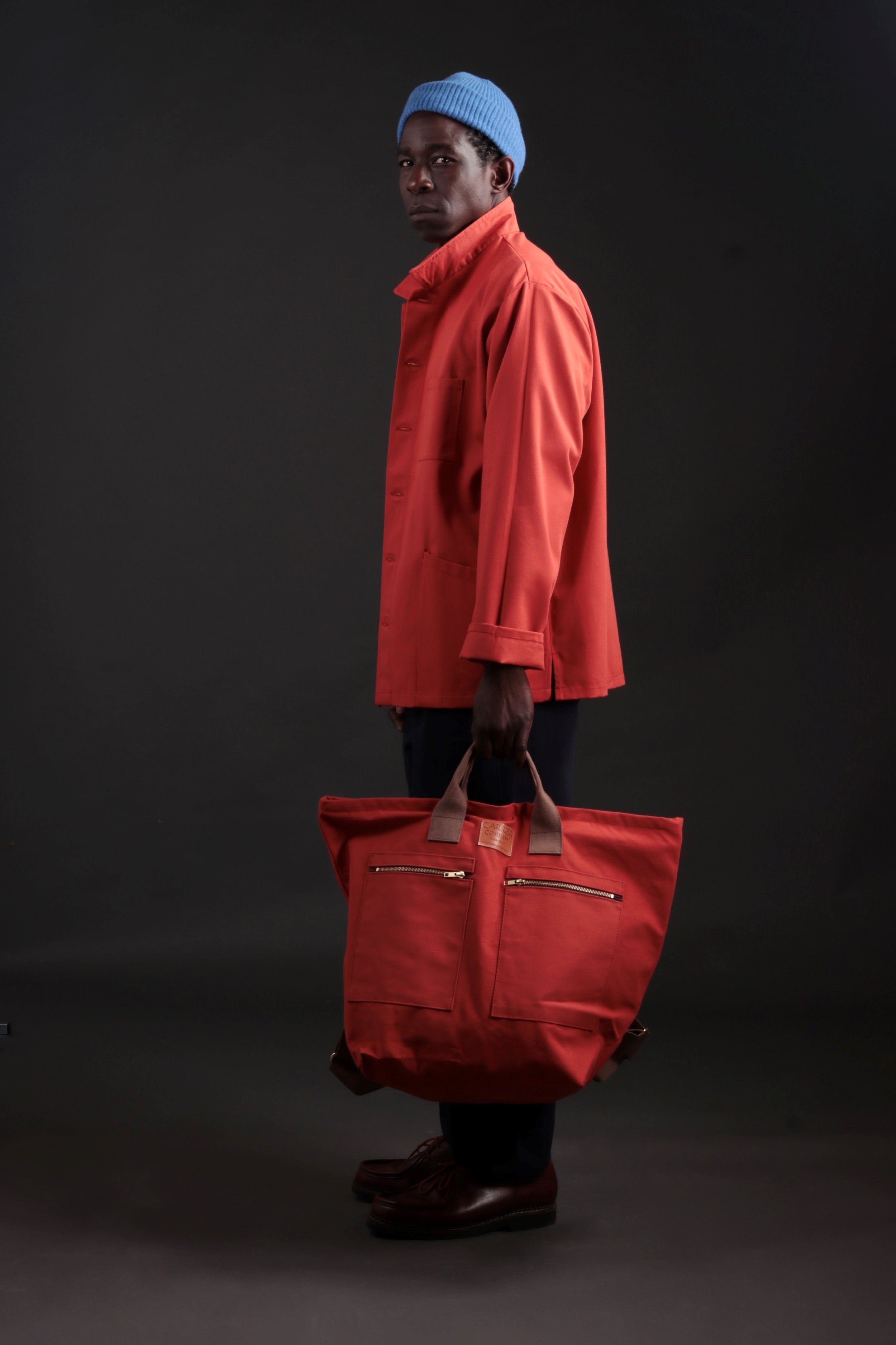 Man wears Carrier Company Norfolk Work Jacket in Orange with a Turquoise Donegal Hat and carries a Backpack in Orange