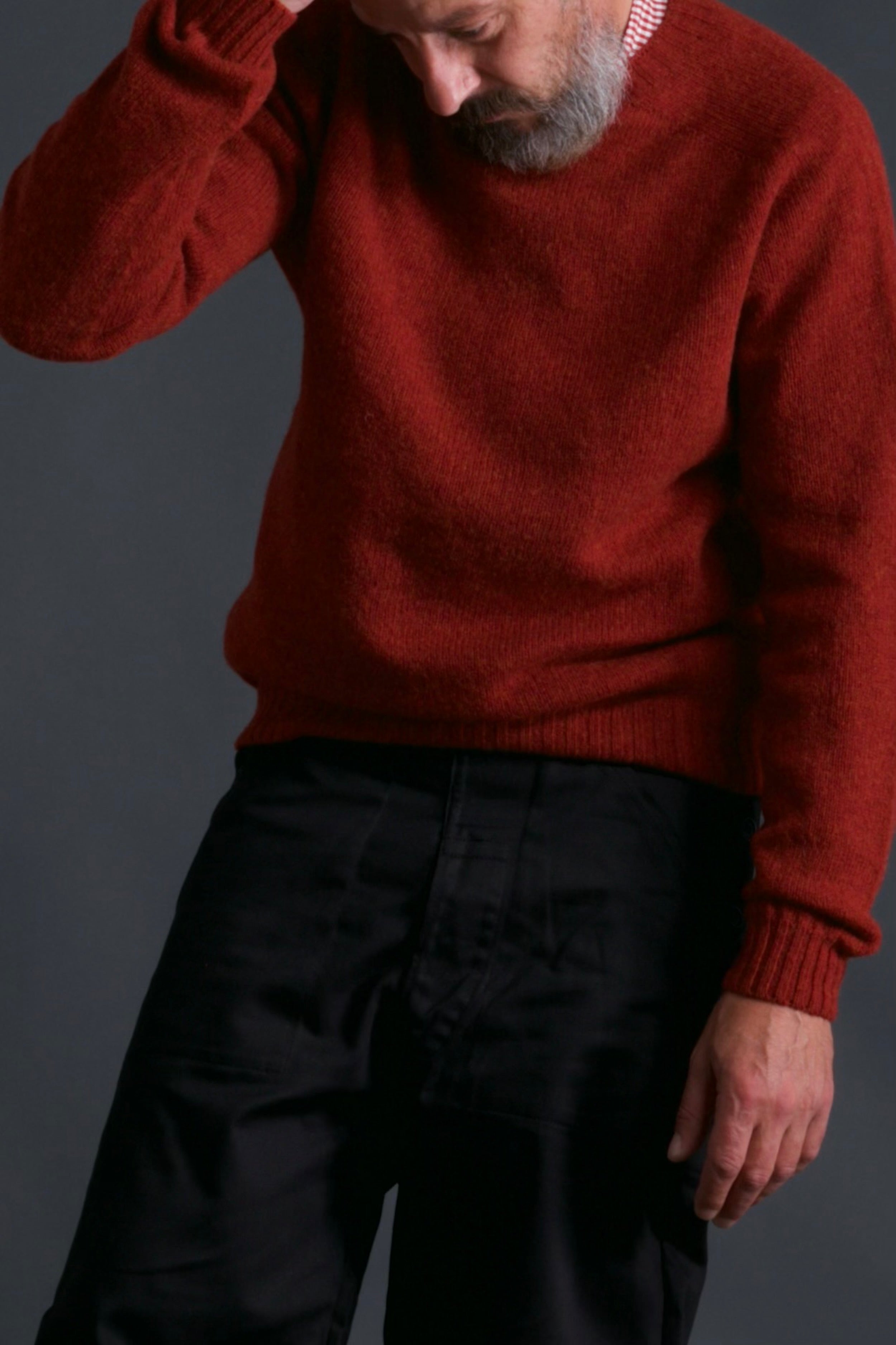 Man wears Carrier Company Shetland Lambswool Jumper in Blaze with Black Work Trousers and Red Gingham Collarless Shirt