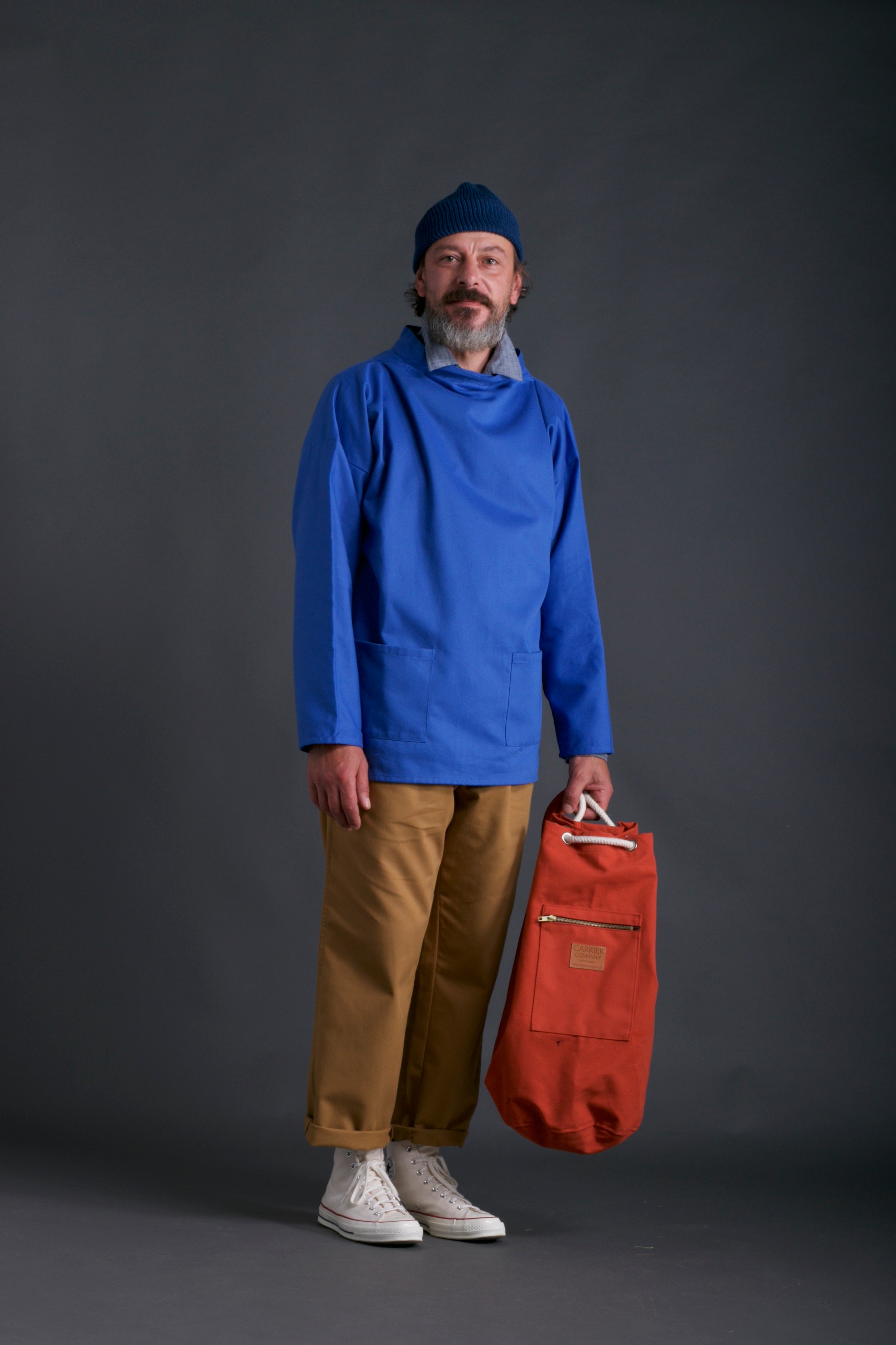 Man wears Carrier Company Traditional Slop in Sky, Classic Trouser, Chambray Shirt, Wool Hat and Carrier Company Duffel Bag in Orange