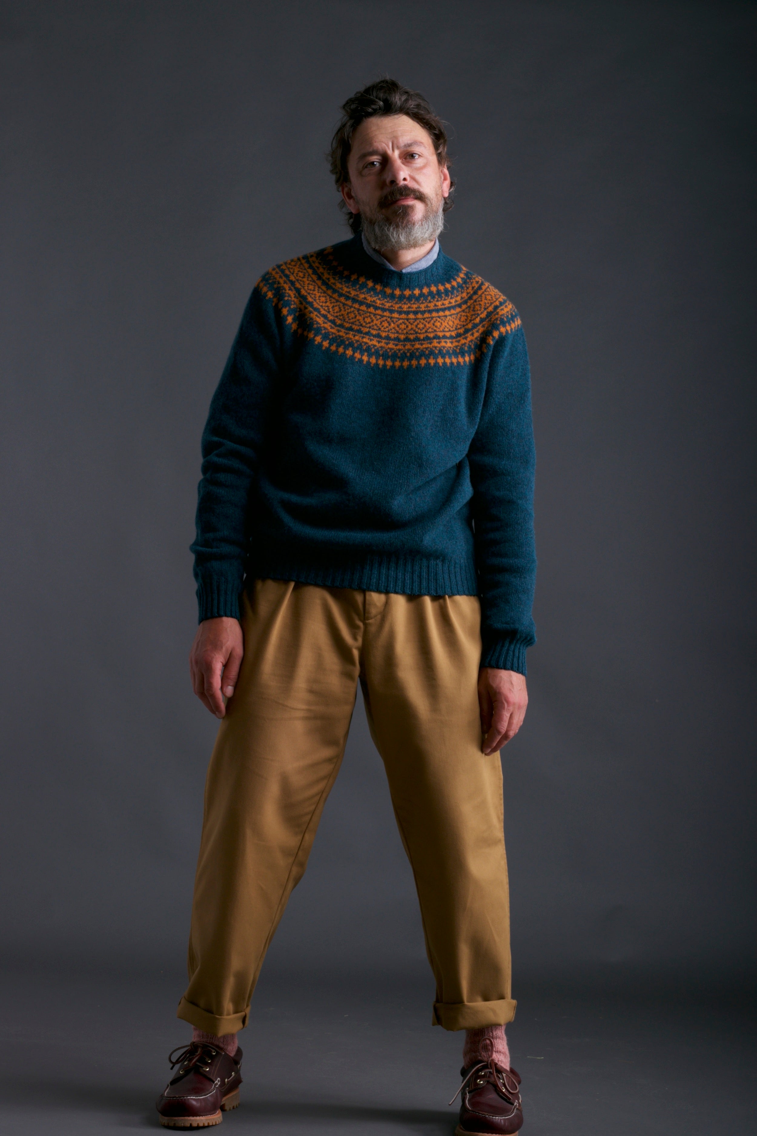 Man wears Carrier Company Shetland Yoke Jumper in ginger and Teal with Mens Work Trouser in Tan