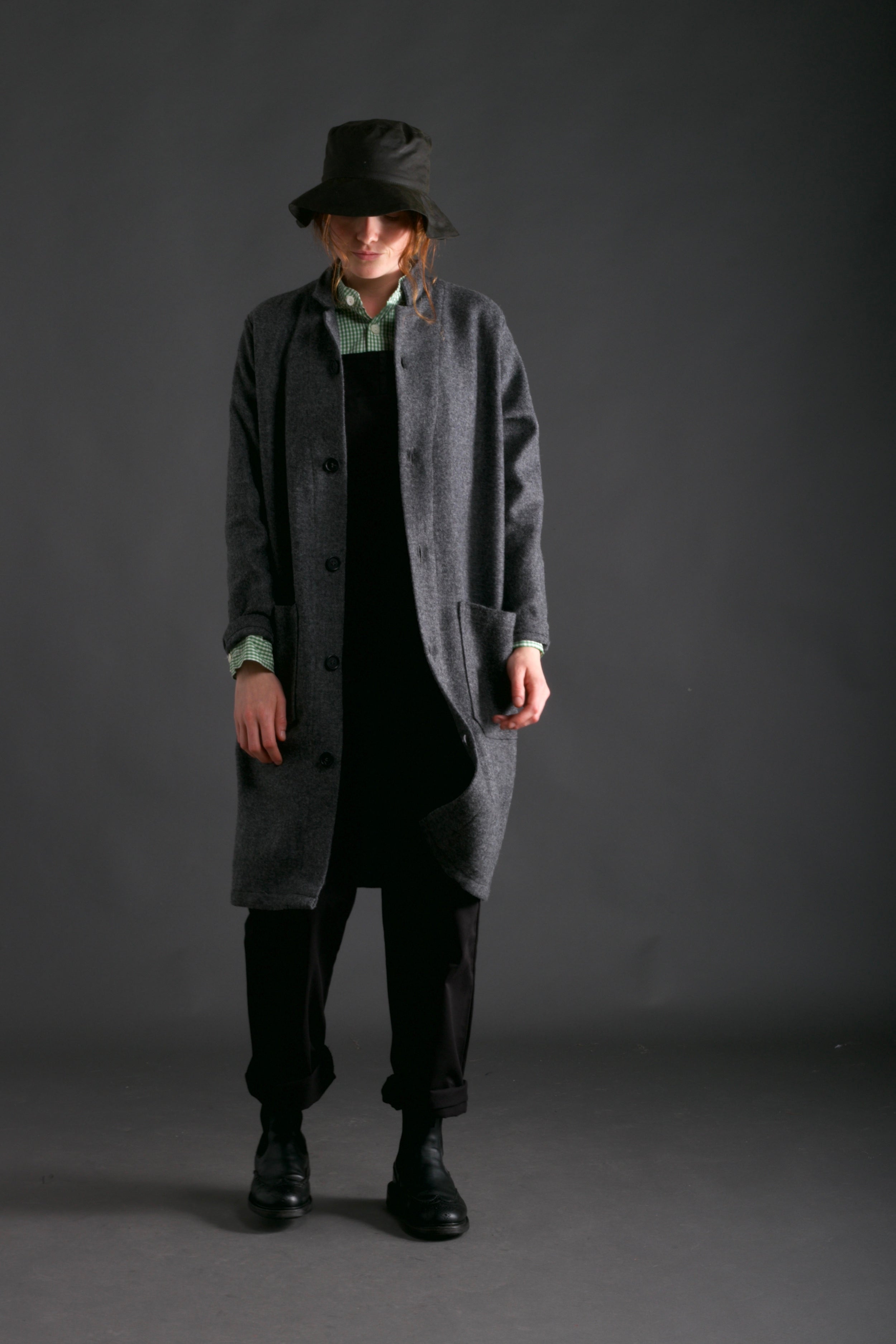 Woman wears Wool Coat in Grey with Women's Dungarees in Black, Gingham Collarless Work Shit and Waxed Cotton Rain Hat in Olive
