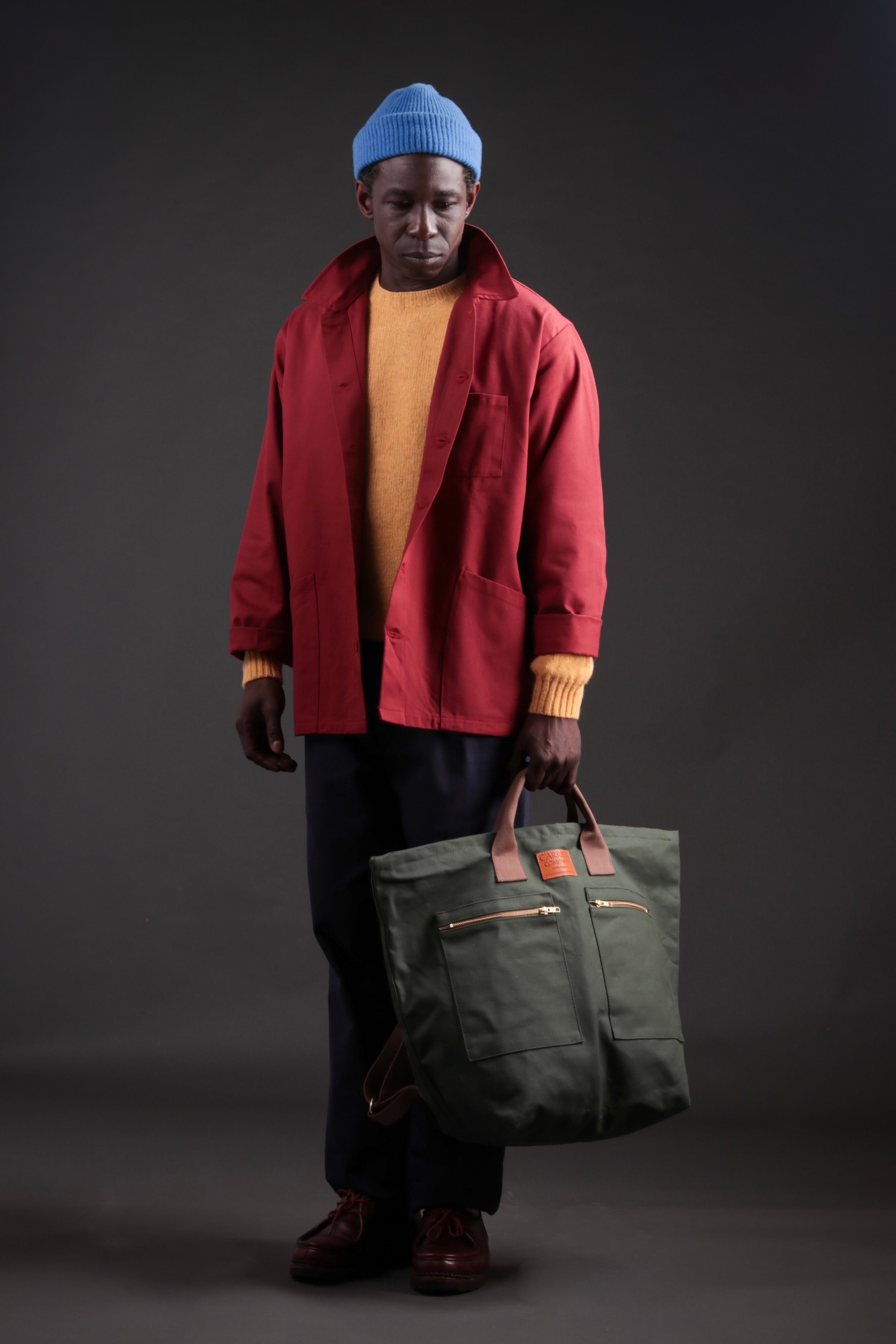 Man wearing Carrier Company Shetland Lambswool Jumper in Chamomile, Breton Norfolk Work Jacket and Navy Classic Trousers. Man is holding Carrier Company Olive Backpack.
