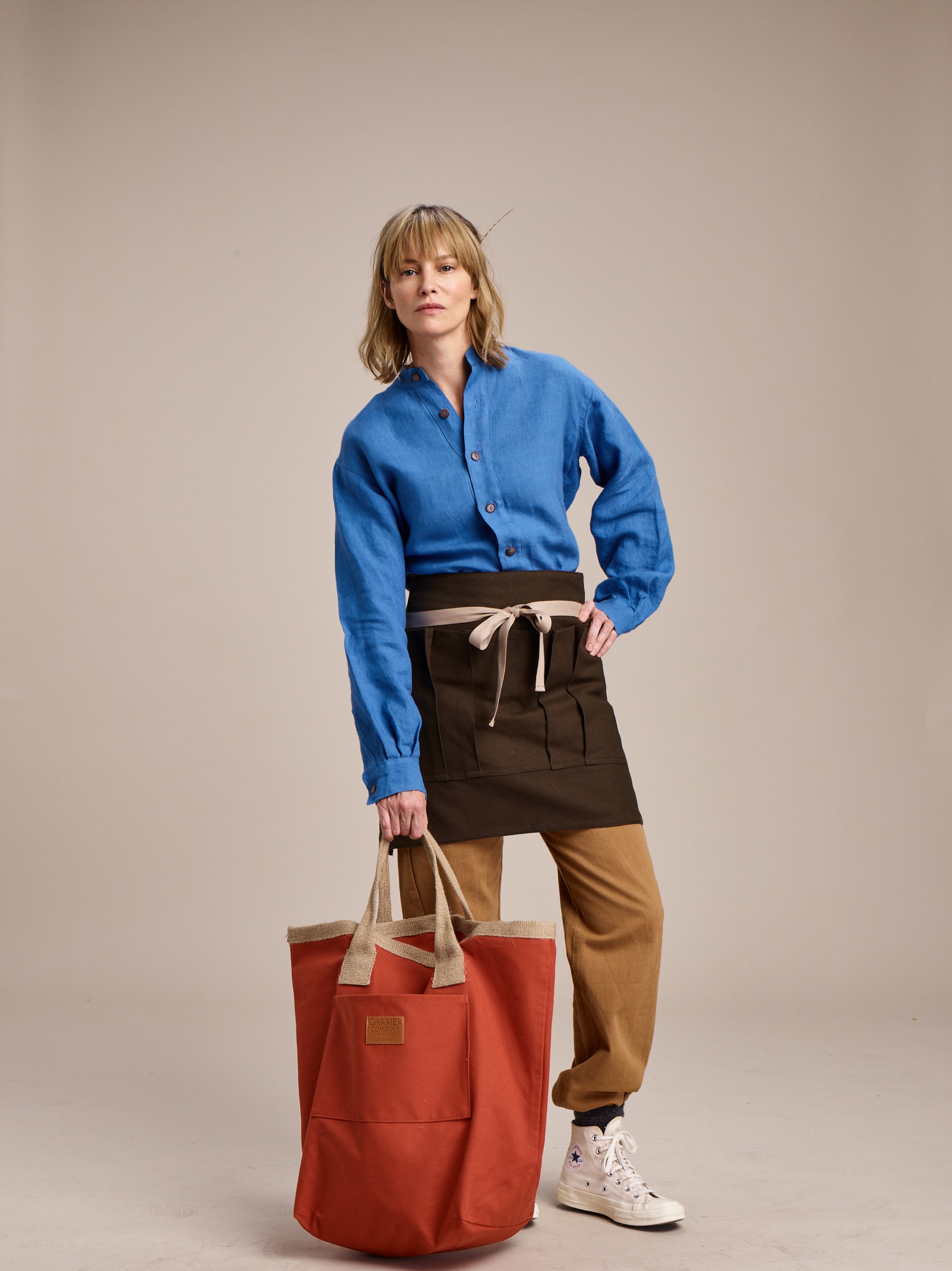 Woman wearing Carrier Company Women's Work Trouser in Tan with Linen Collarless Shirt and Half Apron holding Loot & Boot Bag