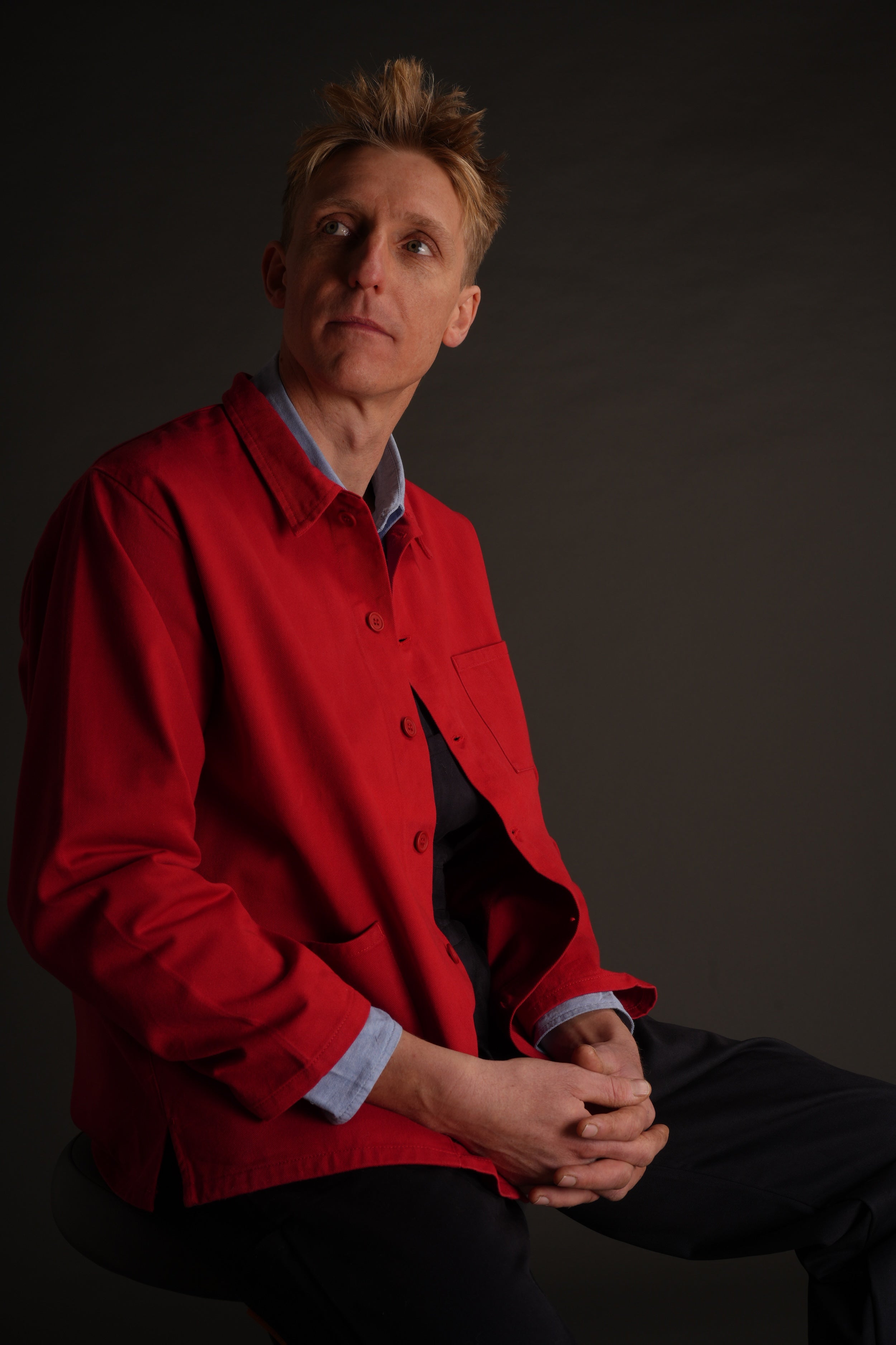 Tommy wears Carrier Company red garment washed cotton work jacket over navy blue dungarees and pale blue chambray shirt with no socks and white plimsoles