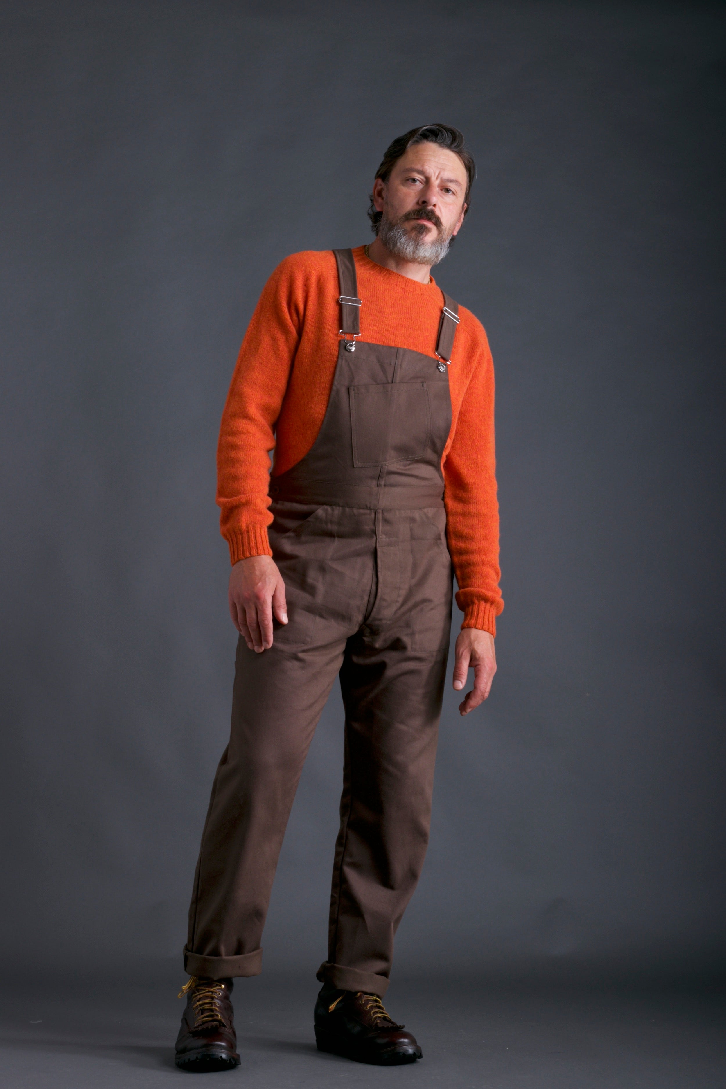 Man wears Carrier Company Men's full-length Dungarees in Olive with Shetland Lambswool Jumper in Tangerine