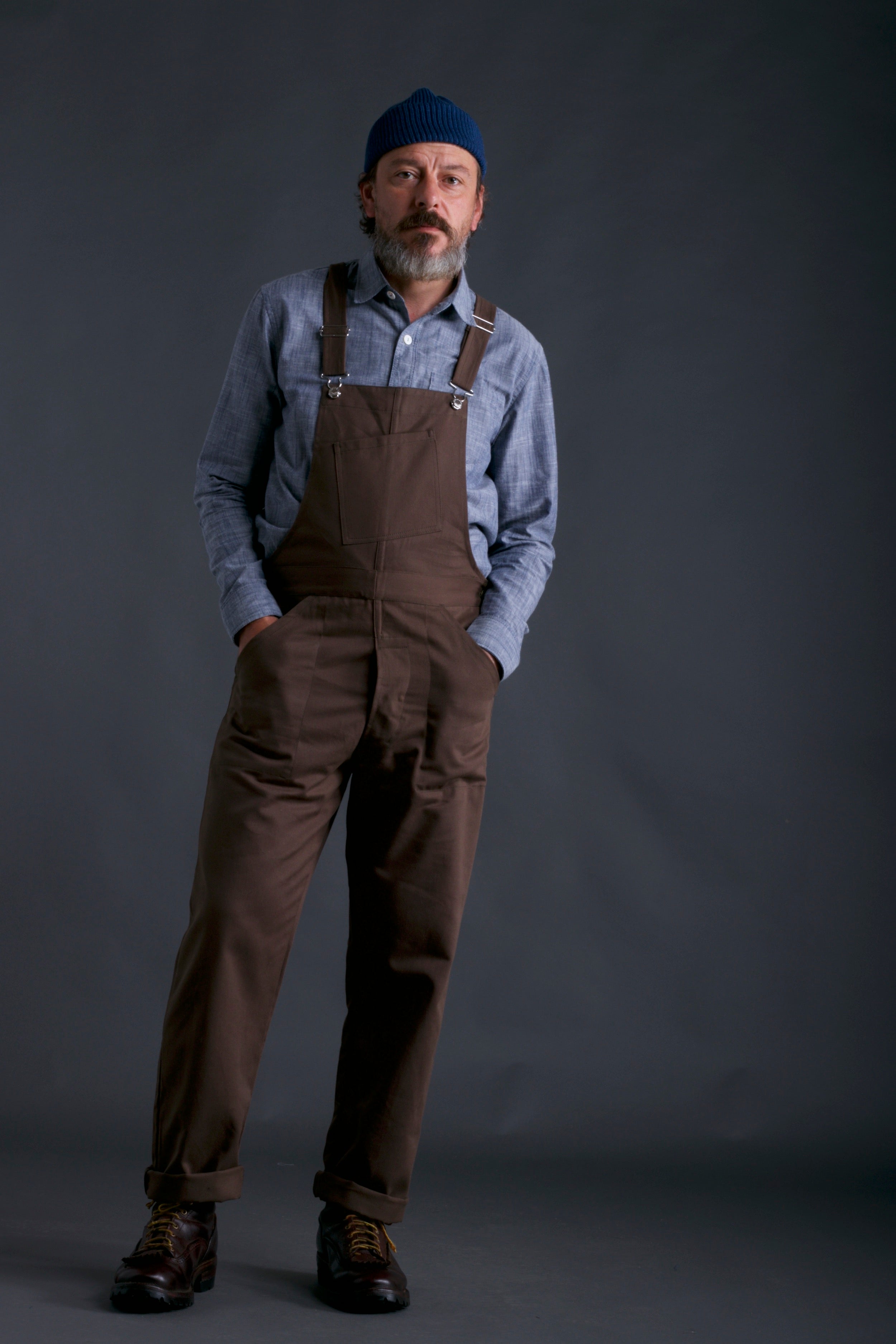 Man wears Carrier Company Men's Dungarees in Olive with Chambray Shirt and Petrol Blue Wool Merino Hat