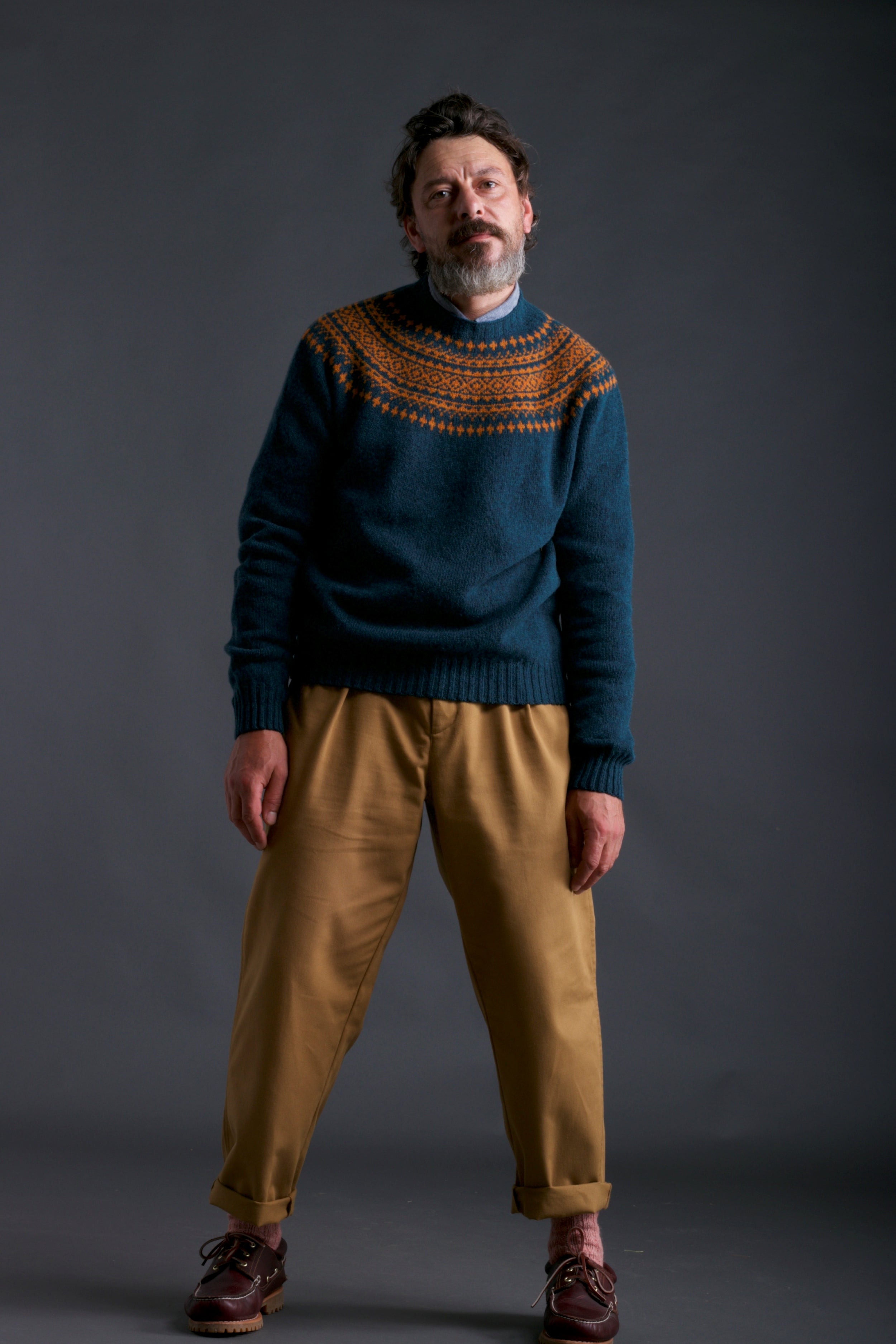 Man wears Carrier Company Classic Trouser in Tan and Shetland Lambswool Yoke Jumper in Ginger & Teal