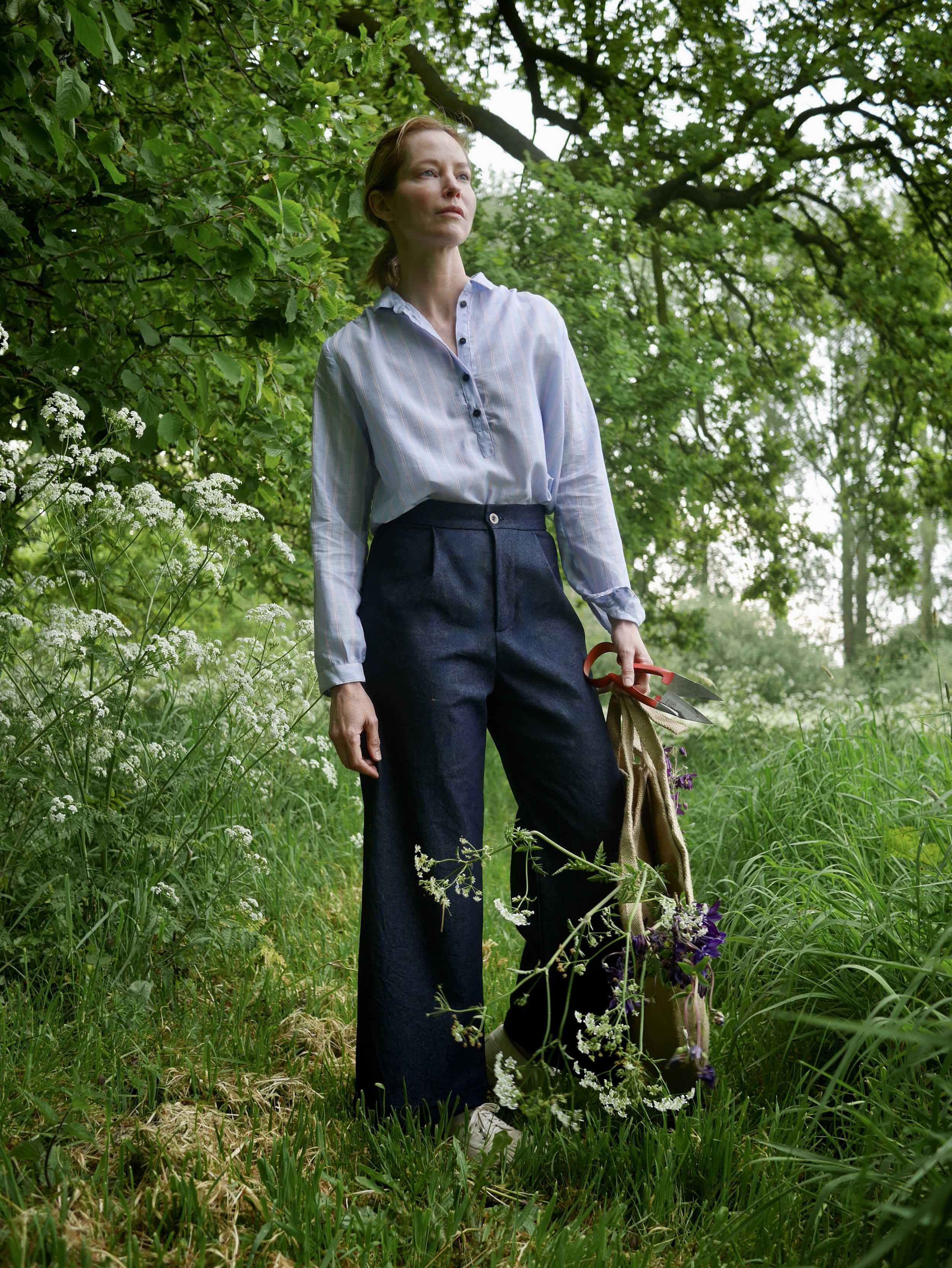 Woman wears sheer striped pullover blouse in blue, and white with a fine red stripe with denim Sailor Trousers and holds the classic carrier