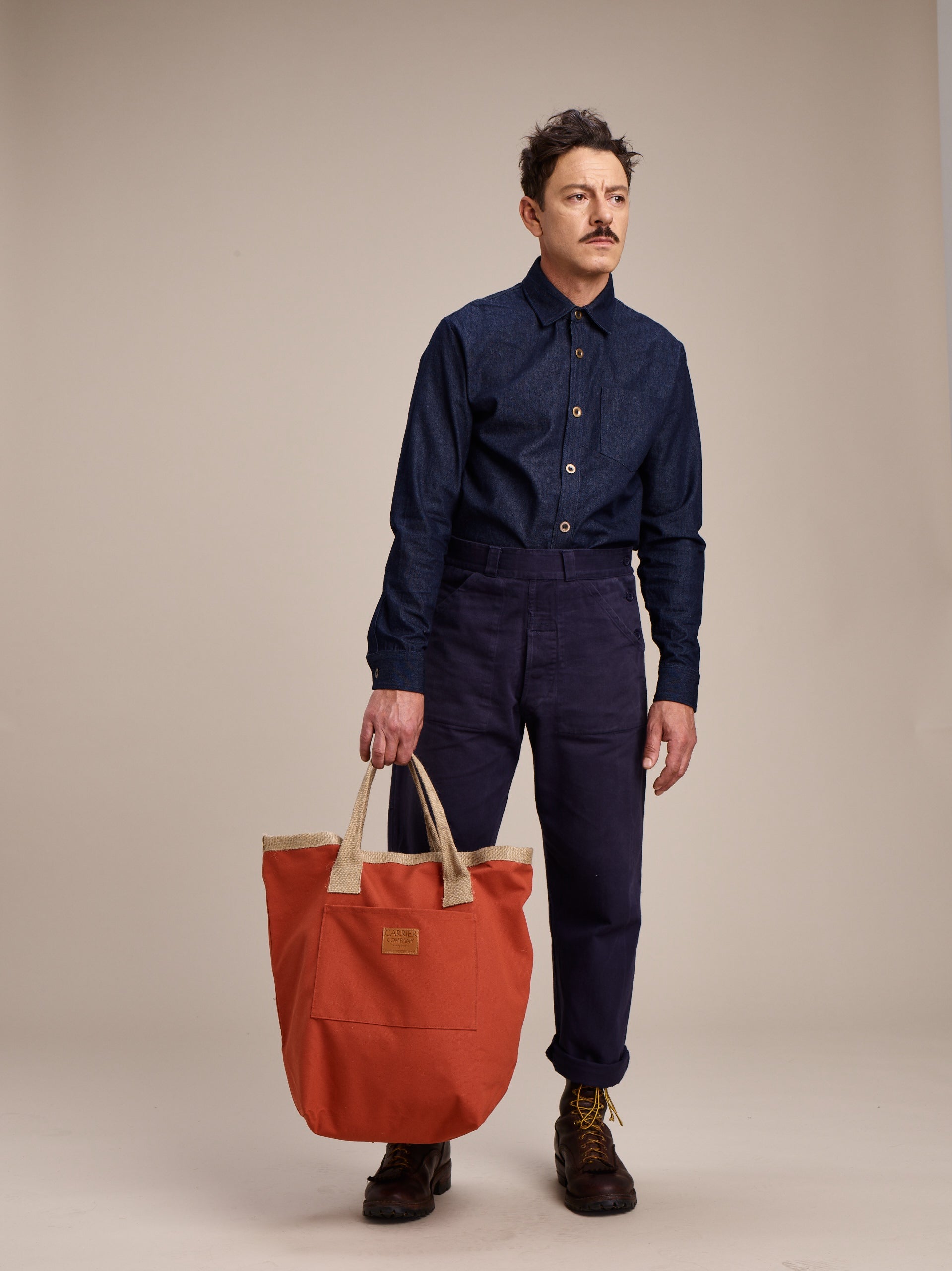 Man Wearing Carrier Company Denim Collar Shirt with Men's Work Trouser in Navy holding Loot & Boot Bag in Orange