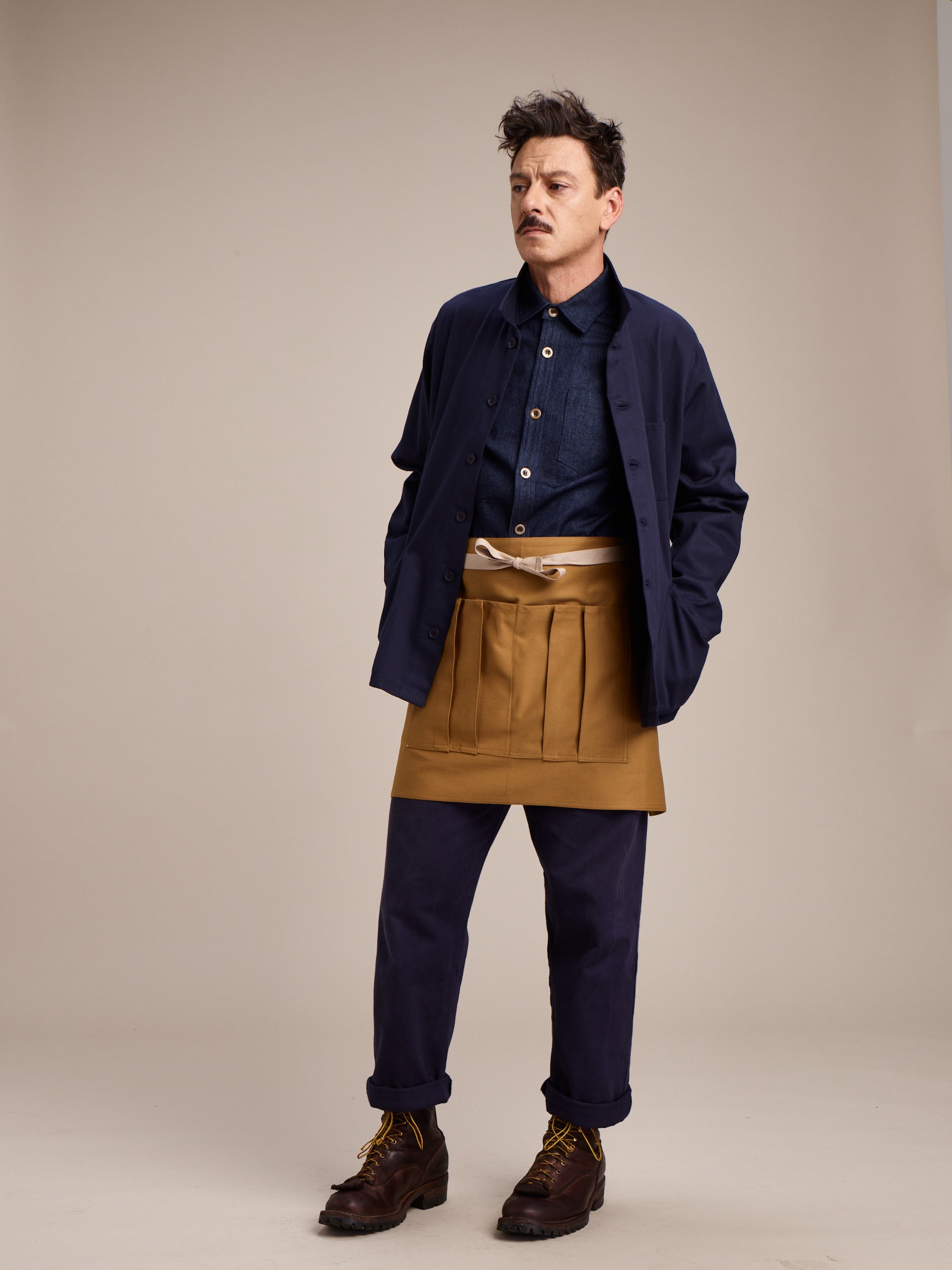 Man wearing Carrier Company Traditional Norfolk Work Jacket in Navy with Denim Collar Shirt and Navy Work Trouser and Gardener's Half Apron