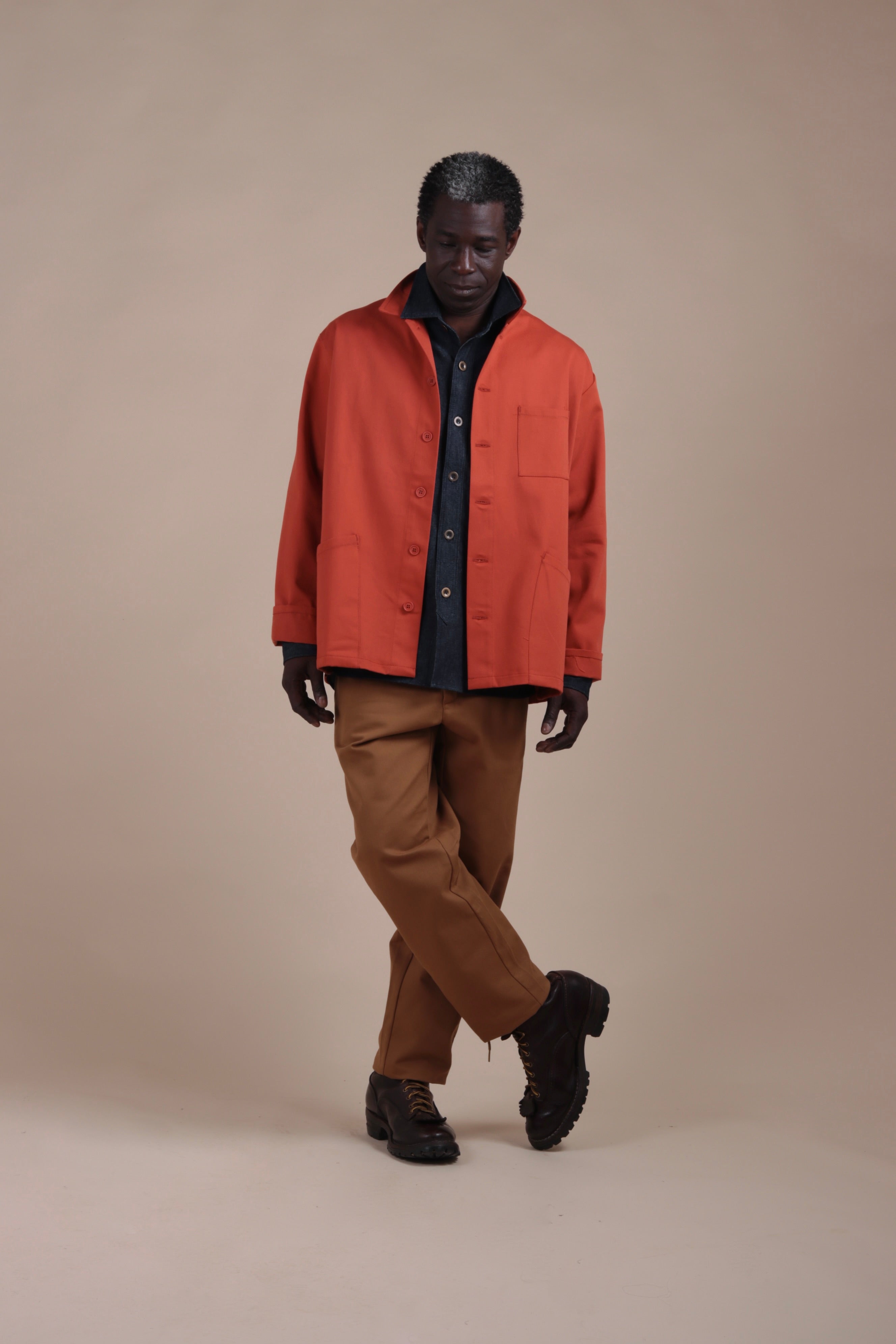 Man wears Carrier Company Norfolk Work Jacket in Orange with Denim Collar Shirt and Classic Trousers in Tan