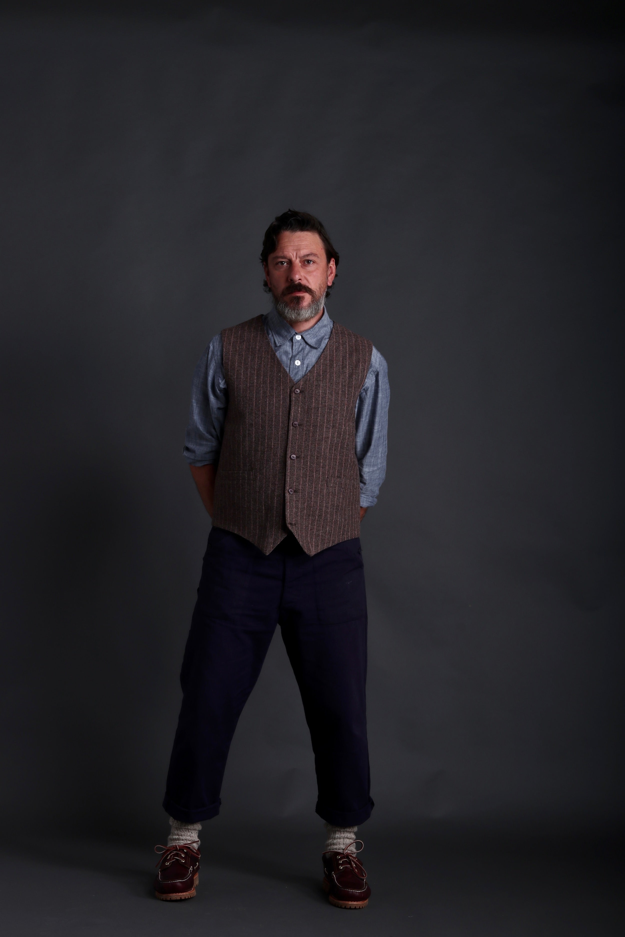 Man wears Carrier Company Men's Wool Waistcoat in Ginger and Pink Herringbone with Chambray Shirt and Classic Trouser