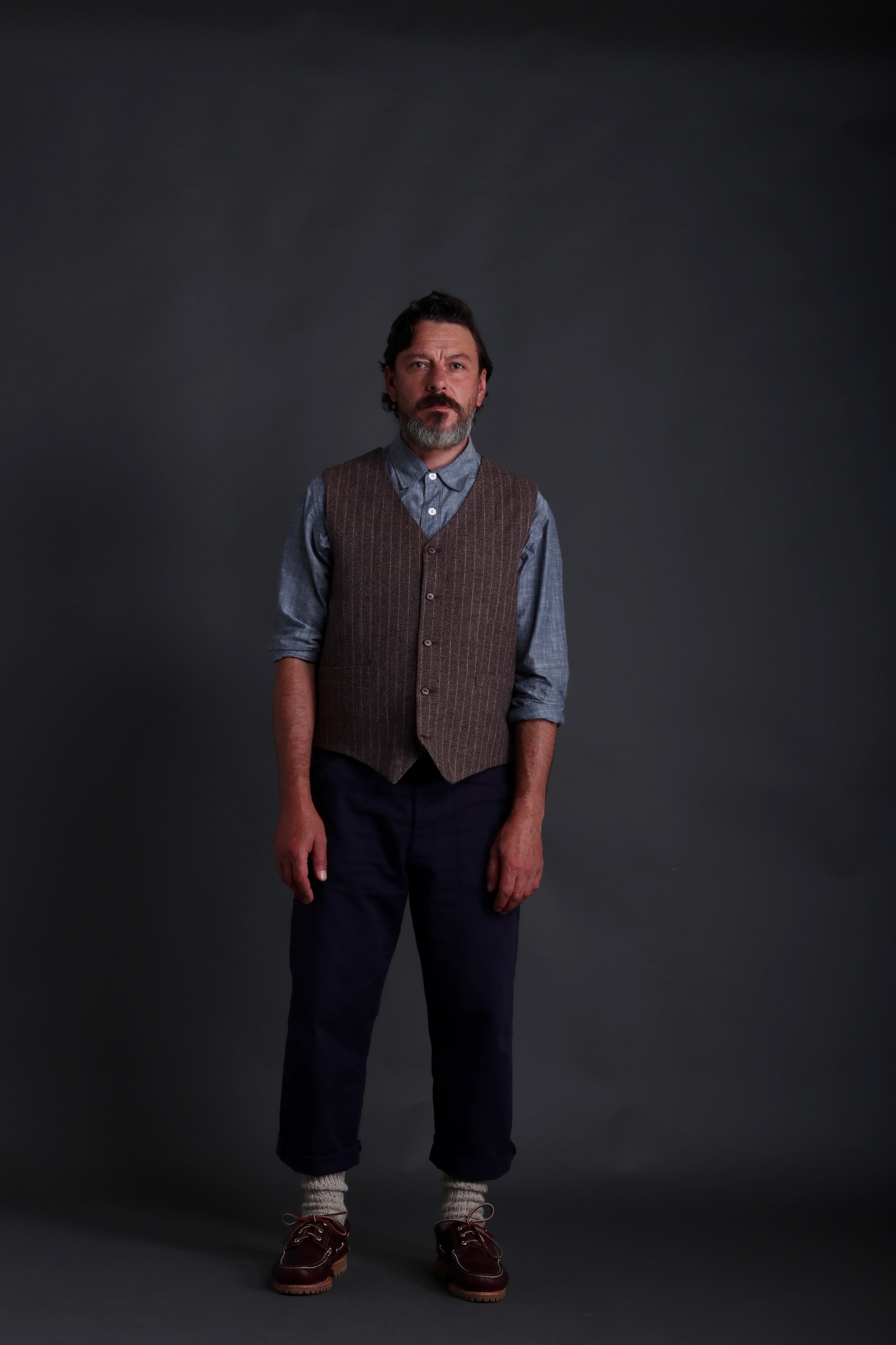 Man wears Carrier Company Men's Wool Waistcoat in Ginger and Pink Herringbone with Chambray Shirt and Classic Trouser