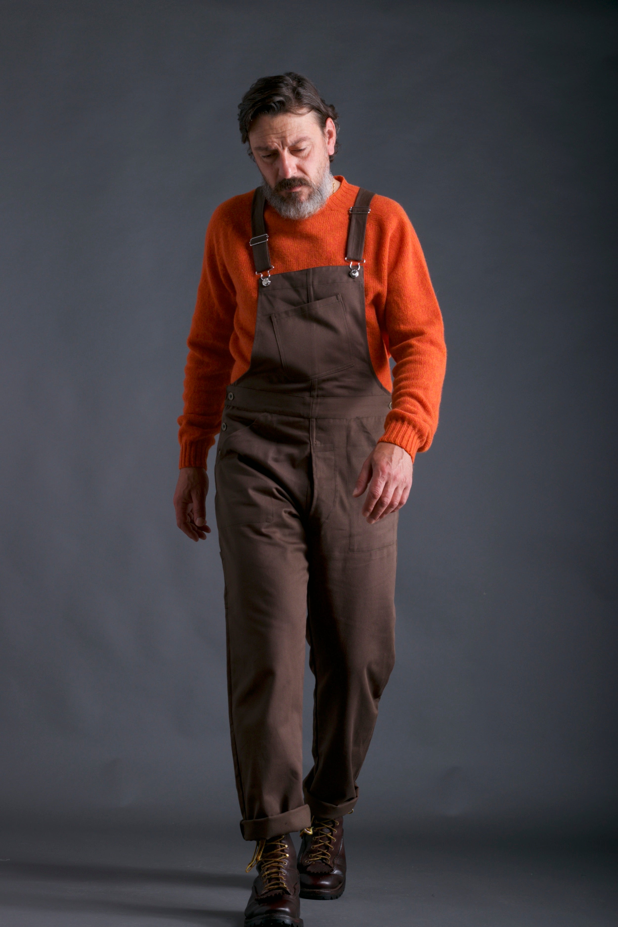 Man wears Carrier Company Men's full-length Dungarees in Olive with Shetland Lambswool Jumper in Tangerine