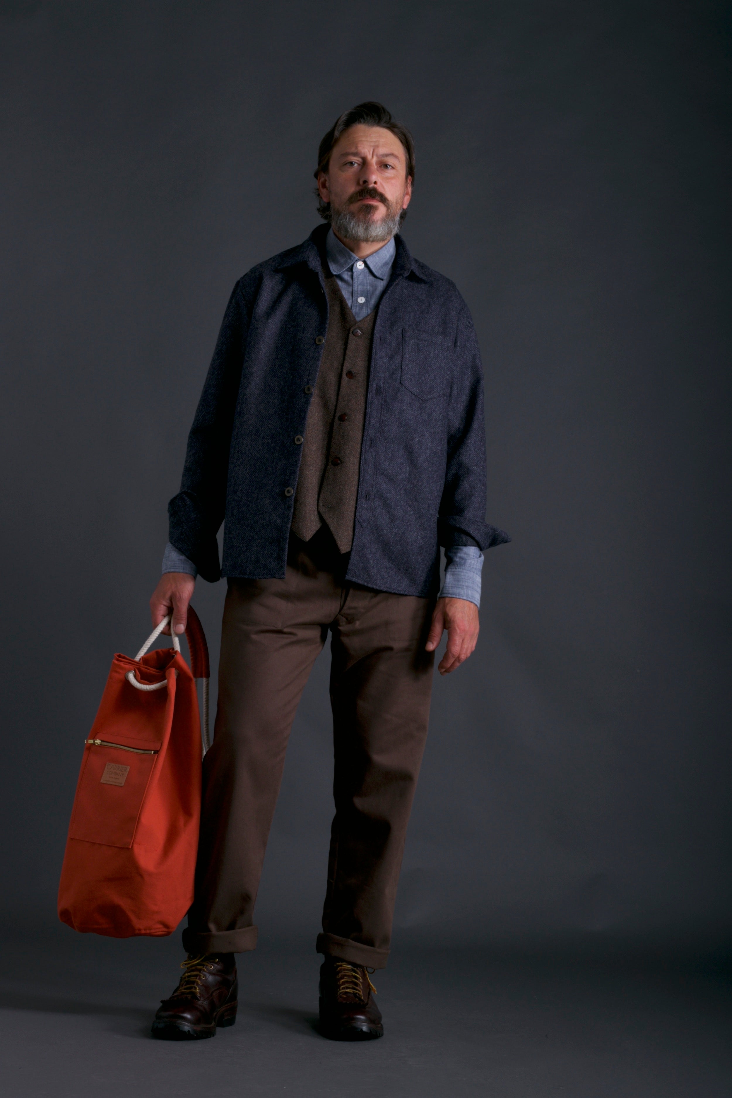 Man wears Carrier Company Wool Overshirt, Wool Waistcoat, Chambray Shirt, Work trouser in Olive and Duffel Bag in Orange