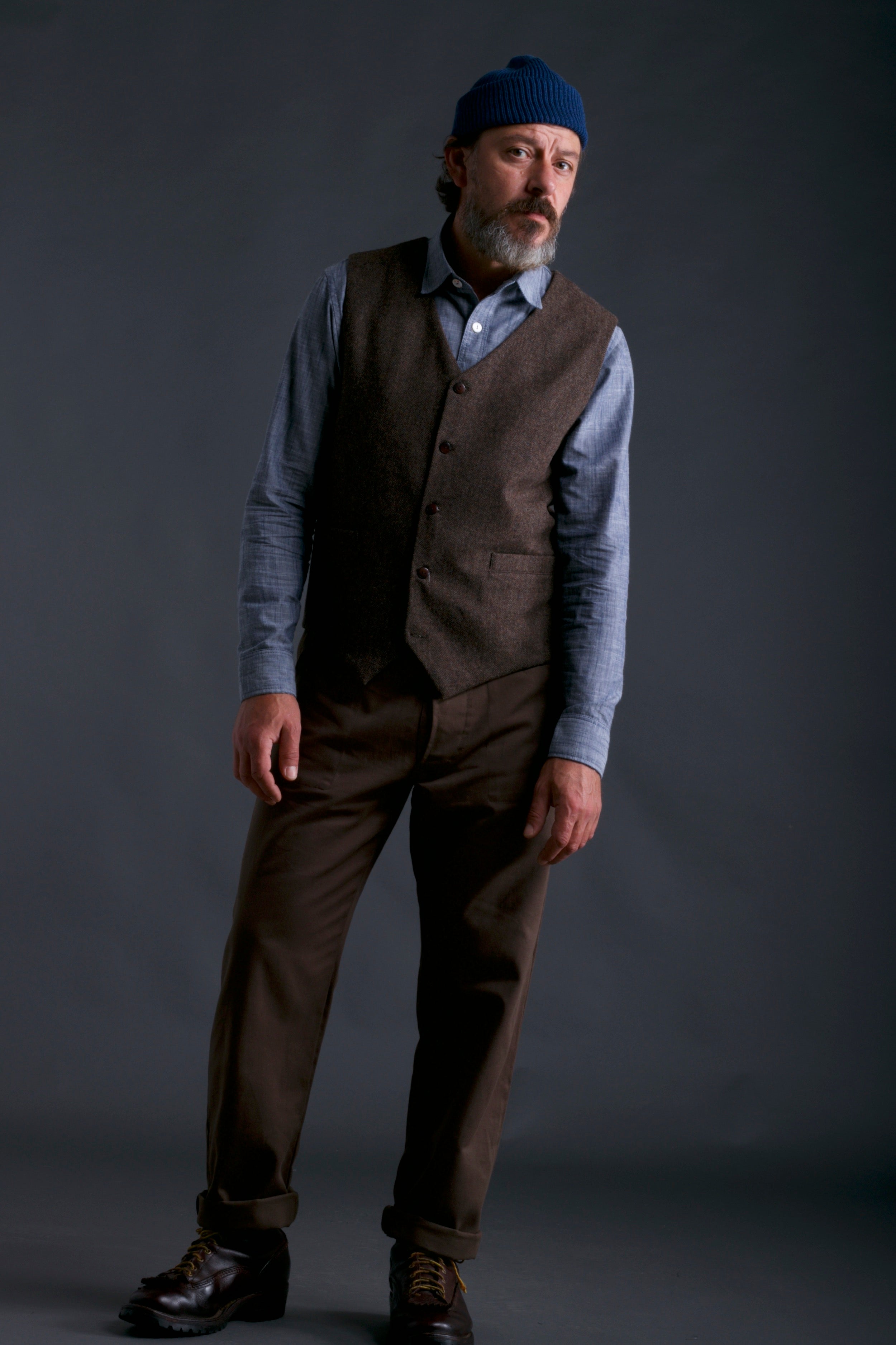 Man wears Carrier Company Men's Wool Waistcoat in Ginger/Brown Herringbone with Chambray Shirt, Wool Hat in Petrol Blue and Olive Dungarees