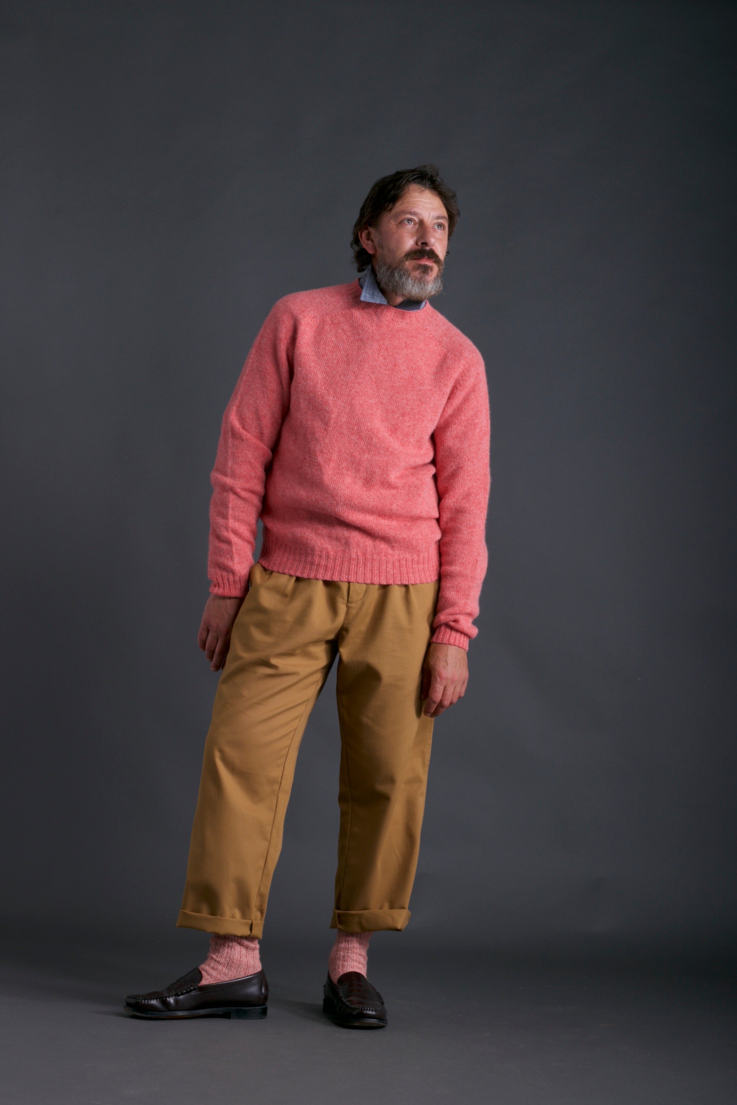Man wears Carrier Company Shetland Lambswool Jumper in Salmon with Chambray Shirt and Classic Trouser in Tan