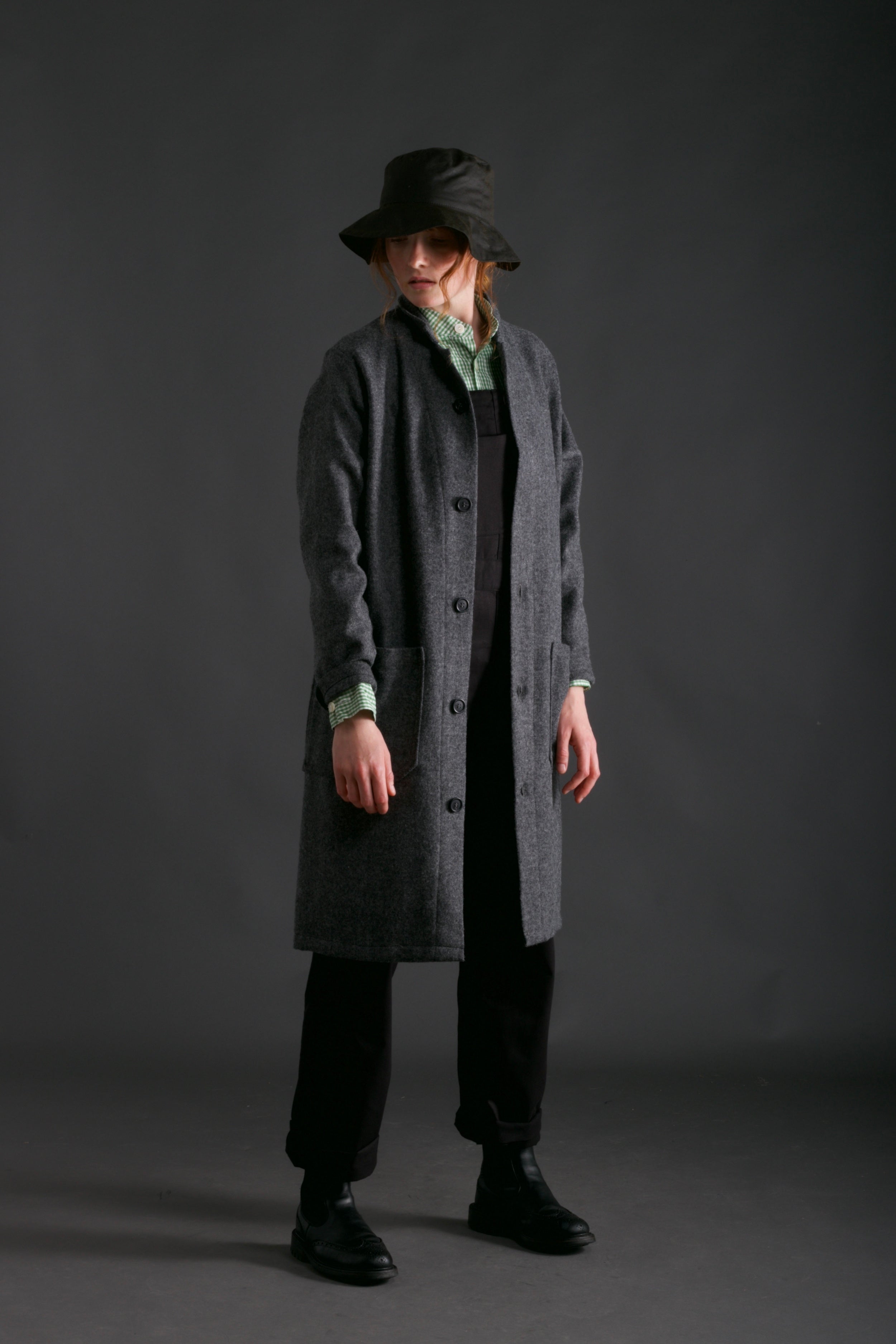 Woman wears Wool Coat in Grey with Women's Dungarees in Black, Gingham Collarless Work Shit and Waxed Cotton Rain Hat in Olive