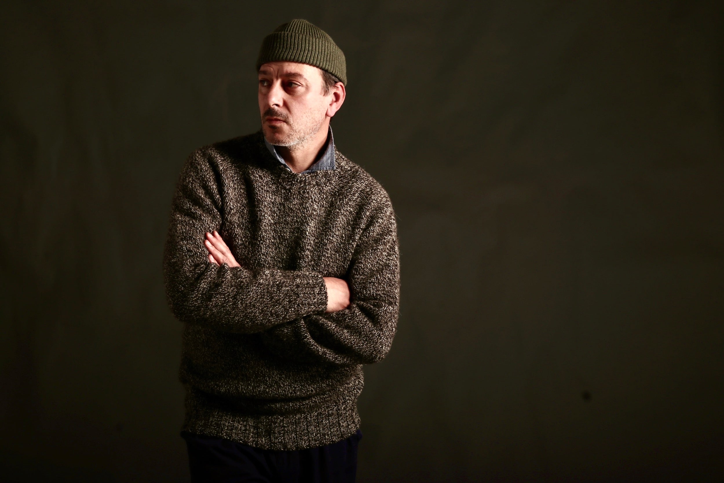 Man wears Carrier Company Heavy Heritage Wool jumper in Jacob, a blend of natural undyed wool ranging from Cream to Tan to Brown to Black, woven together to create an overall dark speckled appearance, Worn with Mens Classic Trouser and Wool Hat in Olive Drab