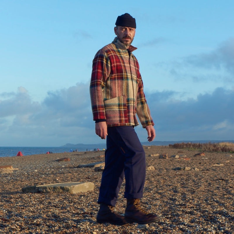 Man wearing the Carrier Company red Celtic Wool Jacket with blue trousers and utility boots on a pebble beach with the sea in the background